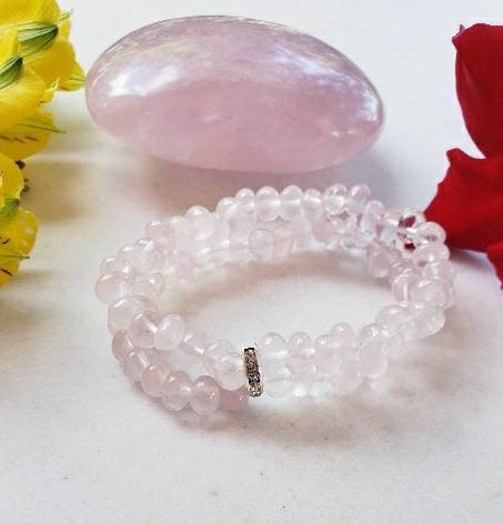 May include: A double-strand bracelet made of light pink rose quartz beads. The beads are smooth and oval-shaped. The bracelet has a silver clasp with small clear stones.