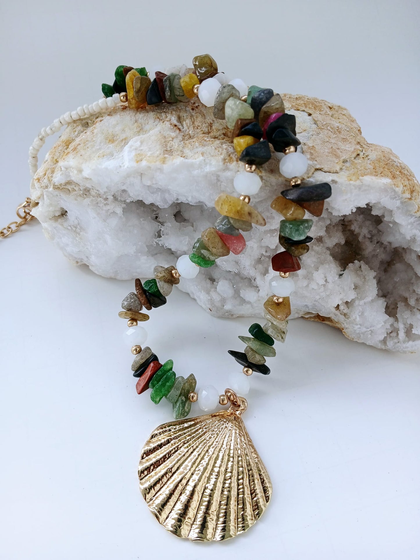 May include: Multicolored gemstone necklace with a gold seashell pendant. The necklace features a mix of small, irregularly shaped stones in shades of green, red, yellow, brown, and black, interspersed with white beads.  The gold pendant is a detailed, textured seashell design. The necklace is displayed on a white geode.