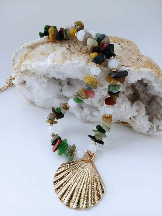 May include: Multicolored gemstone necklace with a gold seashell pendant. The necklace features a mix of small, irregularly shaped stones in shades of green, red, yellow, brown, and black, interspersed with white beads.  The gold pendant is a detailed, textured seashell design. The necklace is displayed on a white geode.