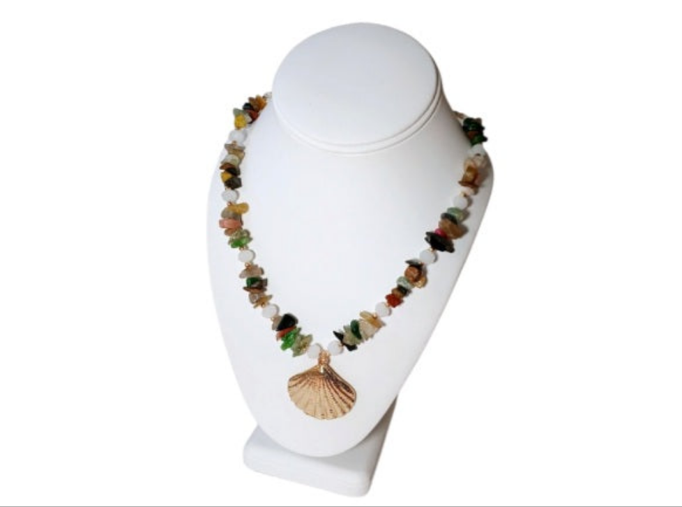 May include: Multicolored gemstone necklace with a gold seashell pendant. The necklace features a mix of small, irregularly shaped stones in shades of green, red, yellow, brown, and black, interspersed with white beads.  The gold pendant is a detailed, textured seashell design. The necklace is displayed on a white geode.