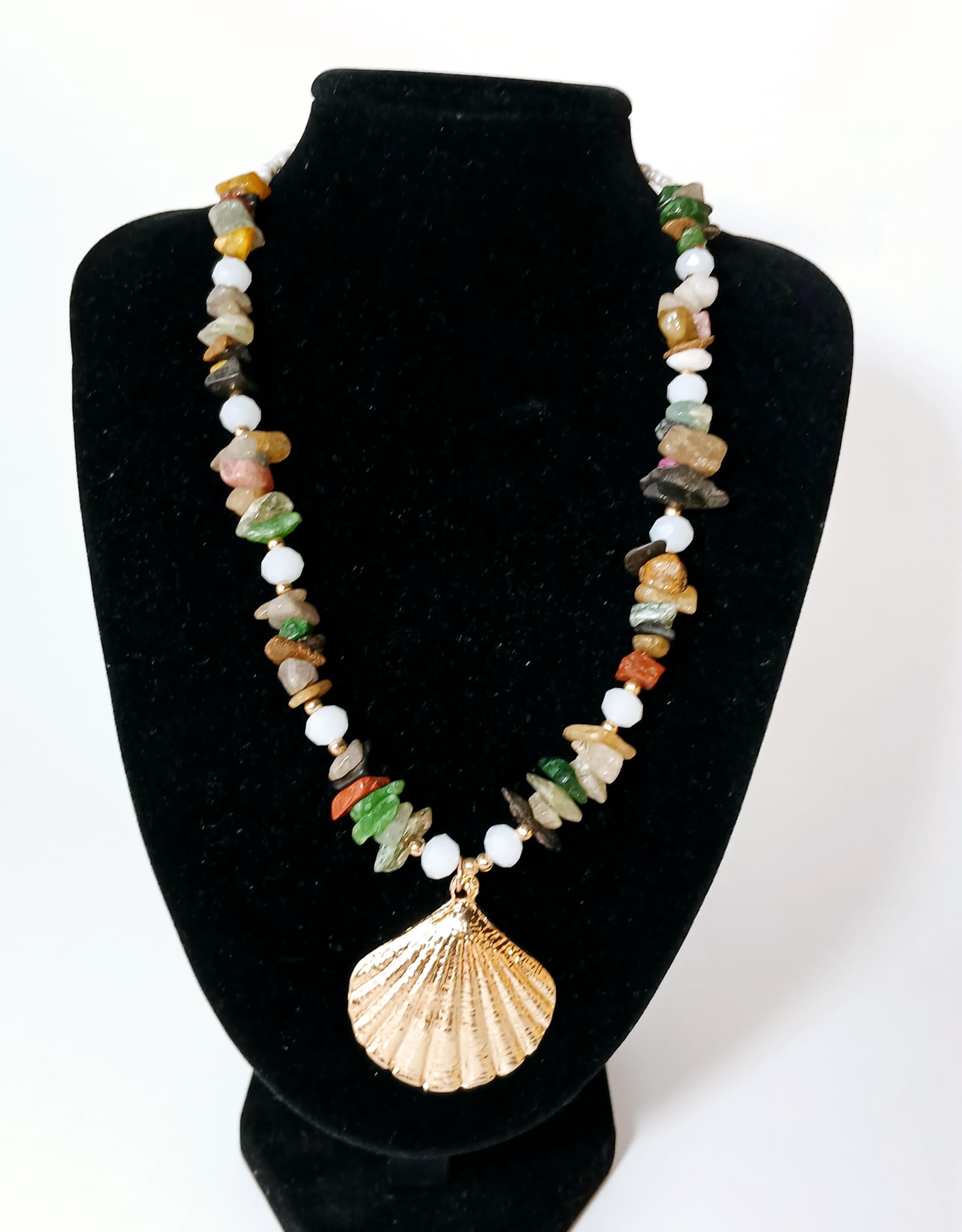 May include: Multicolored gemstone necklace with a gold seashell pendant. The necklace features a mix of small, irregularly shaped stones in shades of green, red, yellow, brown, and black, interspersed with white beads.  The gold pendant is a detailed, textured seashell design. The necklace is displayed on a white geode.
