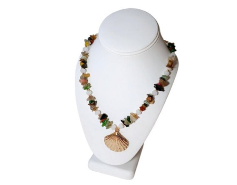 May include: Multicolored gemstone necklace with a gold seashell pendant. The necklace features a mix of small, irregularly shaped stones in shades of green, red, yellow, brown, and black, interspersed with white beads.  The gold pendant is a detailed, textured seashell design. The necklace is displayed on a white geode.