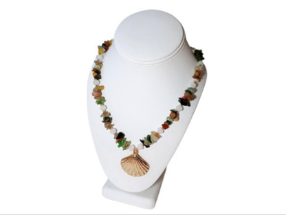 May include: Multicolored gemstone necklace with a gold seashell pendant. The necklace features a mix of small, irregularly shaped stones in shades of green, red, yellow, brown, and black, interspersed with white beads.  The gold pendant is a detailed, textured seashell design. The necklace is displayed on a white geode.