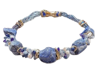 May include: A necklace made of blue and white stones with gold accents. The necklace is made of large, irregular shaped stones. The stones are strung together with gold wire. The necklace is displayed on a white mannequin.