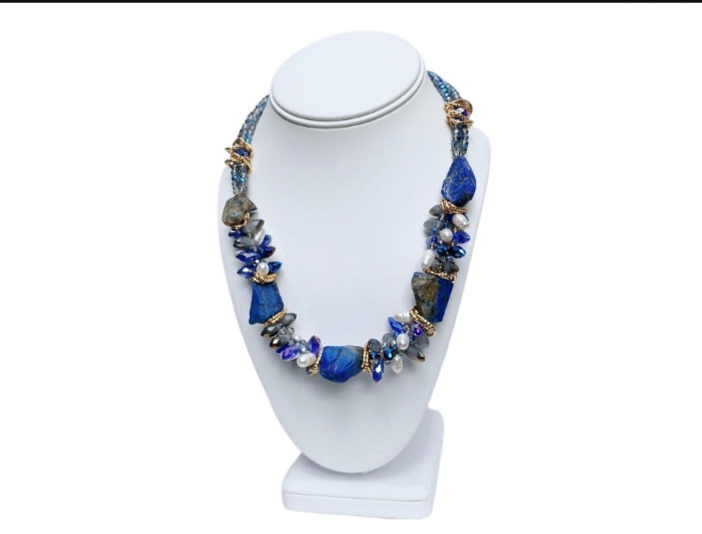 May include: A necklace made of blue and white stones with gold accents. The necklace is made of large, irregular shaped stones. The stones are strung together with gold wire. The necklace is displayed on a white mannequin.