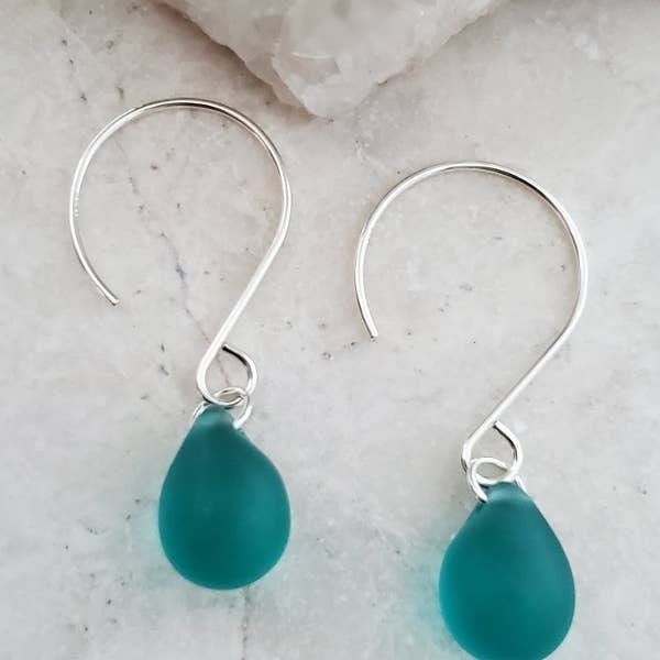 May include: Close-up of a teal sea glass dangle earring. The earring features a single, teardrop-shaped piece of sea glass in a vibrant teal color, suspended from a delicate sterling silver hook. The sea glass is smooth and translucent, with subtle variations in color and texture. The silver hook is simple and elegant, complementing the natural beauty of the sea glass. A ruler is in the background for scale.