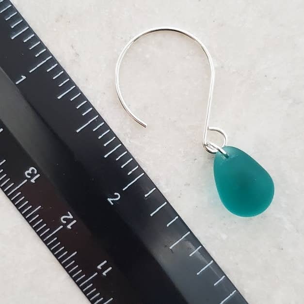 May include: Close-up of a teal sea glass dangle earring. The earring features a single, teardrop-shaped piece of sea glass in a vibrant teal color, suspended from a delicate sterling silver hook. The sea glass is smooth and translucent, with subtle variations in color and texture. The silver hook is simple and elegant, complementing the natural beauty of the sea glass. A ruler is in the background for scale.