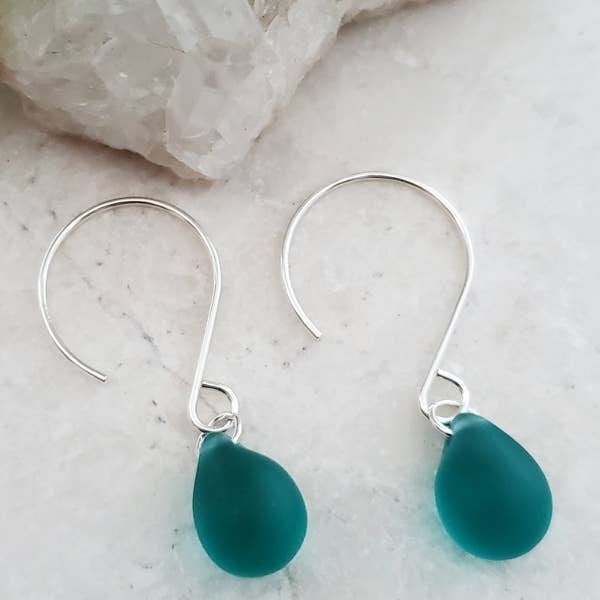 May include: Close-up of a teal sea glass dangle earring. The earring features a single, teardrop-shaped piece of sea glass in a vibrant teal color, suspended from a delicate sterling silver hook. The sea glass is smooth and translucent, with subtle variations in color and texture. The silver hook is simple and elegant, complementing the natural beauty of the sea glass. A ruler is in the background for scale.